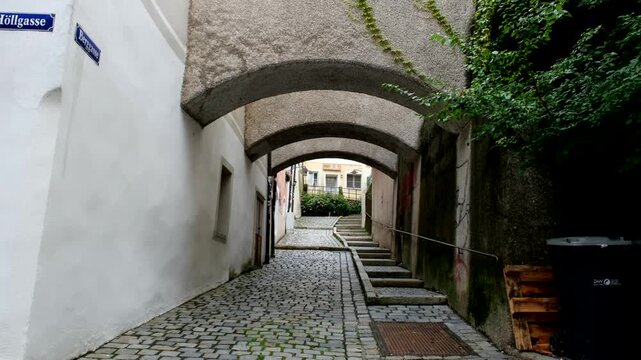 A narrow cobblestone alley with arched ceilings leads to a sunlit courtyard, flanked by old walls and accented with creeping green vines.
