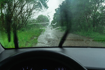 View from inside a car driving on a rainy, muddy road surrounded by green trees, with windshield wipers clearing raindrops for better visibility.