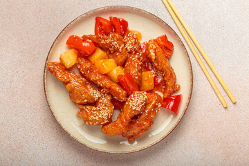 Asian delicious sweet and sour chicken garnished with sesame seeds, pineapple chunks, and bell pepper slices on a plate with chopsticks on light beige background