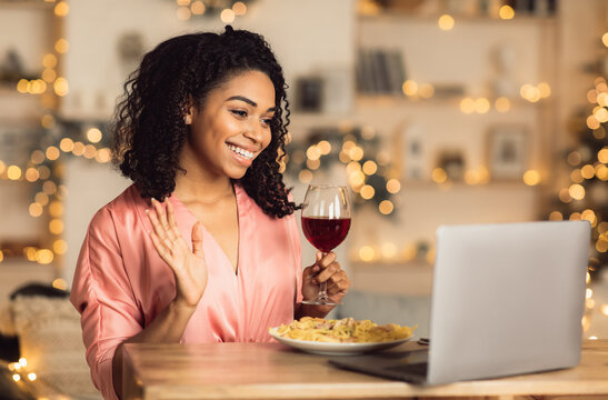 Virtual Wine Testing, Online Event. Smiling cheerful african american woman having video chat with friends or boyfriend during dinner, holding glass, waving hello to webcam, eating pasta