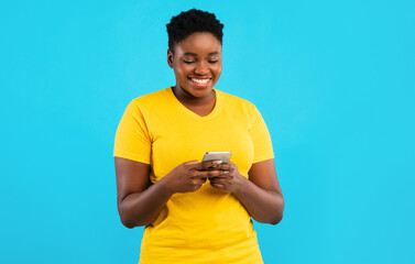 Smiling Black Oversized Lady Using Smartphone Texting Standing Over Blue Studio Background. African...