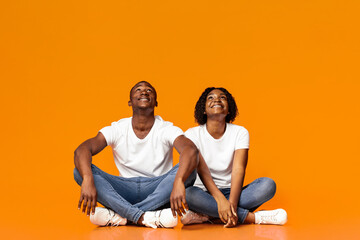 Smiling young black man and woman sitting on floor and looking together upwards at copy space for...