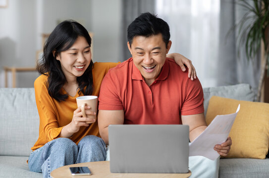 Smiling asian spouses paying bills online while staying home, sitting on couch, using laptop, happy chinese man holding papers and banking on Internet, cheerful woman drinking tea, copy space
