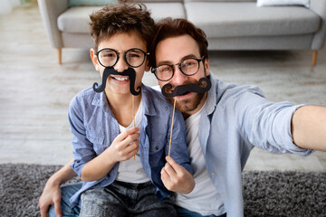 Funny selfie. Happy father and son taking selfie, holding fake moustache on sticks, wearing glasses and smiling at camera, having fun at home, sitting on floor carpet. Parenthood concept