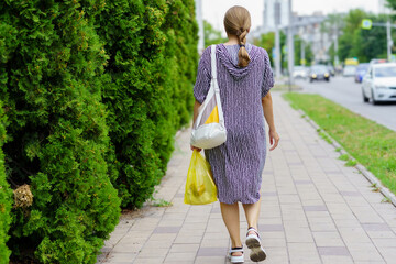 Woman walking on city sidewalk carrying grocery bag in summer attire with lush greenery lining the...