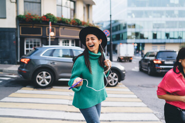 Brunette woman, 30, laughs while listening to music through smartphone and earphones, expressing...