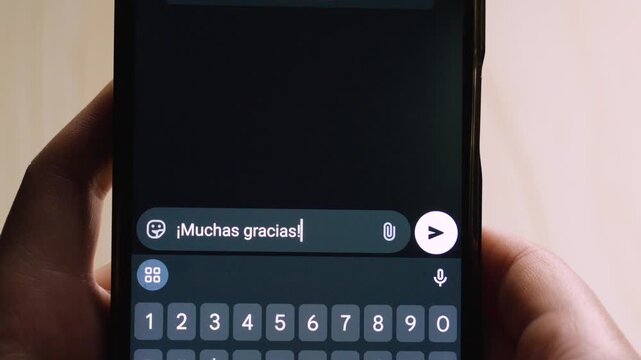 Close-up of a hand holding a smartphone, showing a messaging app with the phrase "&iexcl;Muchas gracias!" typed in Spanish.