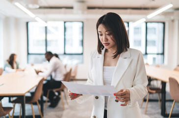 Asian businesswoman reading printed report while standing in bright open office, reviewing charts...