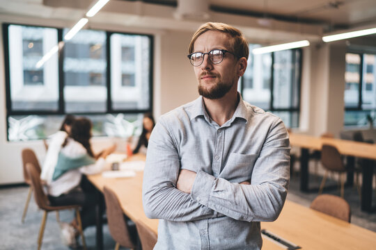 Young thoughtful businessman with folded arms standing in bright office, gazing seriously at distance, contemplating business strategy, decision or problem, team meeting in background