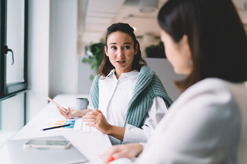 Two stylish young women having casual business conversation by window. Friendly facial expressions, digital tablet, coffee cups and neon sign add visual interest