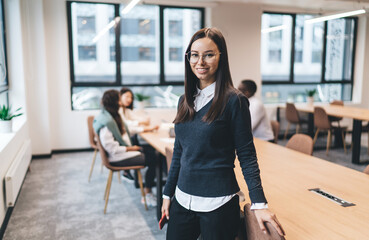 Young businesswoman in glasses standing beside desk, confidently smiling at camera, posing in...