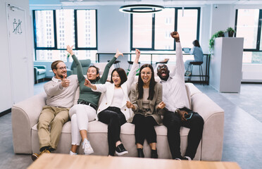 Happy multicultural team celebrating together on couch, cheering with raised hands like fans at...