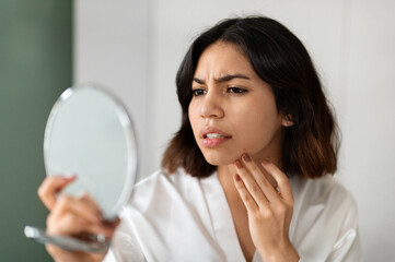 Headshot of upset young arab woman in white silky bathrobe looking at mirror and touching her face...