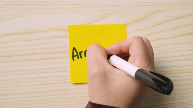 Handwriting the Italian word &ldquo;Arrivederci&rdquo; with a smiley face on a yellow sticky note placed on a wooden desk.