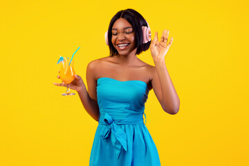 Young black lady in stylish outfit holding exotic cocktail and enjoying favorite music in headphones on orange background. Posh African American woman with tropical drink listening to summer songs