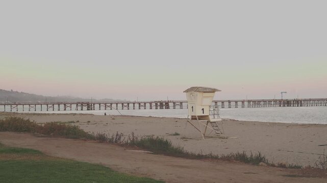 Lifeguard tower and wooden pier on empty beach at dusk with hills behind