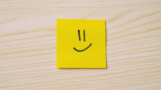 Person using a black marker to draw a smiley face on a yellow sticky note, placed on a wooden desk.
