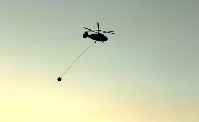 A firefighting helicopter carrying water to extinguish a pine forest fire in the evening of a hot summer day