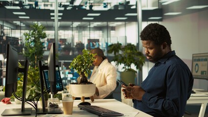 Accountant in modern office using mobile phone to text friends after inputting data on computer. Employee at work typing on smartphone while finishing tasks on PC, camera B