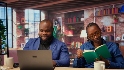 Young woman reading a book next to her boyfriend working remotely, freelancers enjoying their time at their cozy home. African american self employed boyfriend and girlfriend. Camera A.