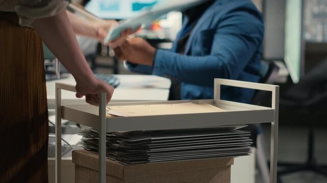 Professional archivist uses rolling trolley to transport documents and records, assisting the ceo and coworkers with efficient handling of critical paperwork in a modern workplace. Camera B.