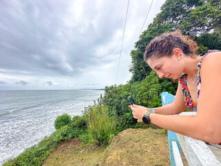 Colombian woman leaning on a fence taking a photo of the sea with her phone from a coastal cliff...
