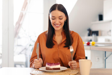 Excited woman getting ready to eat cheesecake on plate, holding fork and knife, sitting at table in...