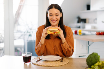Hungry lady holding big burger and smiling, enjoy eating fast food and having cheat meal at home, sitting in kitchen interior