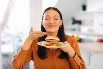 Satisfied woman enjoy fast food snack, smelling burger with closed eyes, being very hungry after hard working day, sitting in kitchen. Eating concept