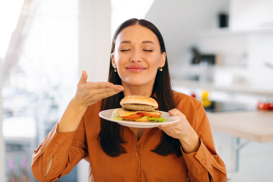Satisfied woman enjoy fast food snack, smelling burger with closed eyes, being very hungry after hard working day, sitting in kitchen. Eating concept