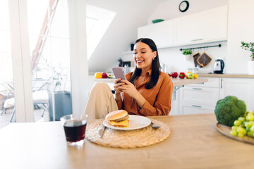 Happy woman using cellphone while eating burger, counting calories or writing feedback for fast...