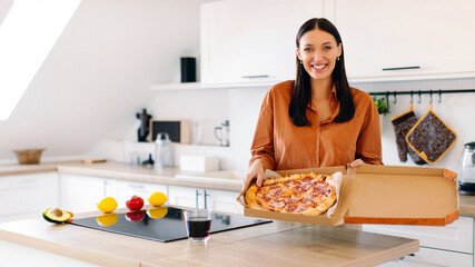 Happy European lady holding open takeaway box with pizza, looking and smiling at camera, standing...