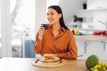 Satisfied lady drinking juice while eating tasty burger at home, sitting at table in kitchen, enjoying takeaway fast food delivery, copy space