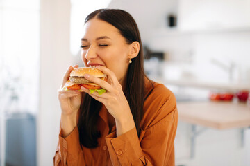 Cheat meal. Woman eating tasty burger, biting delicious fast food snack, sitting in kitchen interior at home, free space