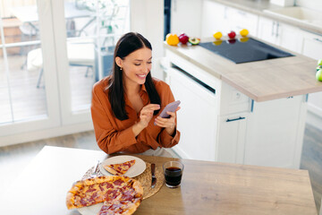 Happy lady using smartphone while eating pizza, writing feedback to delivery service, sitting at table in kitchen, free space, above view