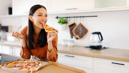 Joyful lady eating pizza alone, biting slice and enjoying taste, satisfied with food delivery, standing at kitchen counter at home