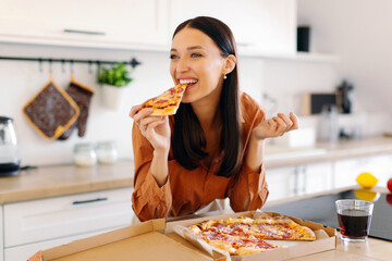 Joyful lady eating pizza alone, biting slice and enjoying taste, satisfied with food delivery, standing at kitchen counter at home