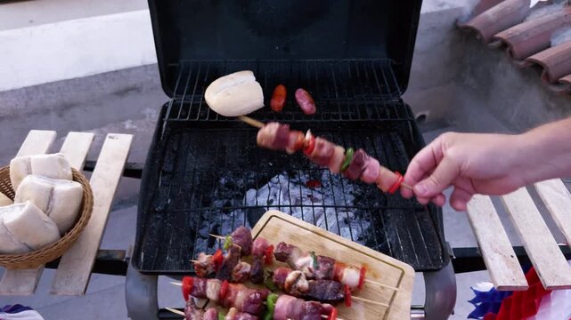 Chilean Independence Day video: Man placing anticuchos from wooden board onto grill with marraquetas and longanizas, seen from terrace