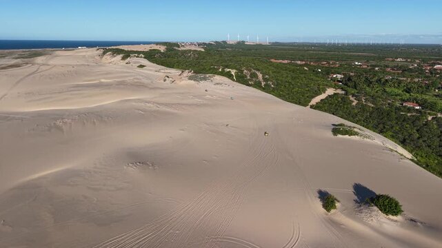 Buggies are driving across the Canoa Quebrada dunes, and wind turbines can be seen in the distance