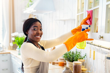 Happy black lady cleaning kitchen cabinet, her husband washing dishes on background. Young couple doing domestic work together, helping each other with house cleanup