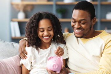 Black girl and father putting coin into piggy bank, sitting on sofa at home. Happy african american dad teaching his cute daughter how to save money, living room interior. Financial education concept