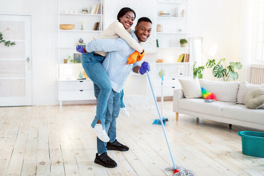 Positive black guy giving piggyback ride to his wife while washing floor at their house, copy space. Young African American couple having fun together, enjoying home cleanup - Powered by Adobe
