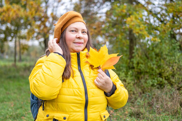 Happy mature woman in beret enjoying autumn nature, with fall leaf in hand, graceful aging, vibrant...
