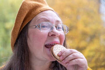 Mature Hungry woman biting sandwich cookies with funny craving expression, comic face expression craving sweet snack, outdoors in fall season, healthy aging
