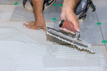 Worker uses trowel to spread adhesive while laying tiles on construction site in broad work day