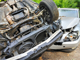 Flipped car is leaning against another vehicle in wooded area following collision, extensive damage.
