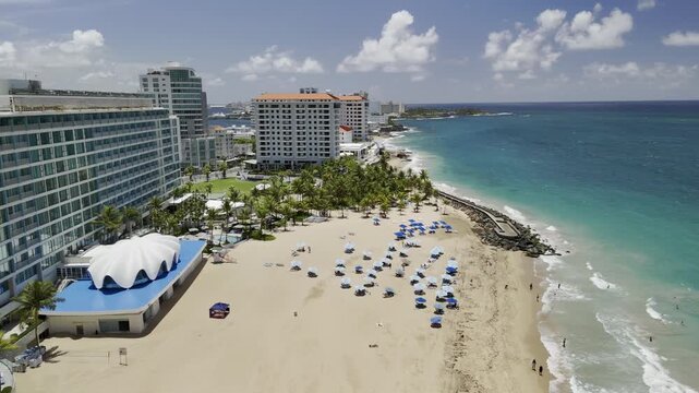 Drone orbits to the right over blue beach umbrellas on sandy beach next to La Ventana al Mar park on sunny morning in San Juan, Puerto Rico
