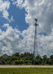 Cell phone tower with antennas along a two lane road near a tree line and old barn with blue sky and fluffy white clouds