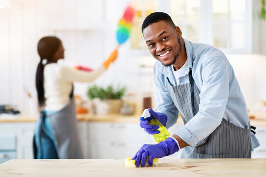 Handsome black guy washing kitchen table while his girlfriend dusting on background, free space. Young African American couple doing domestic duties together, keeping their apartment clean