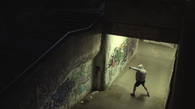 Crane shot of a lone boxer practicing in a graffiti-covered urban underpass. Dim lighting highlights his focused determination and resilience, emphasizing perseverance and inner strength.
